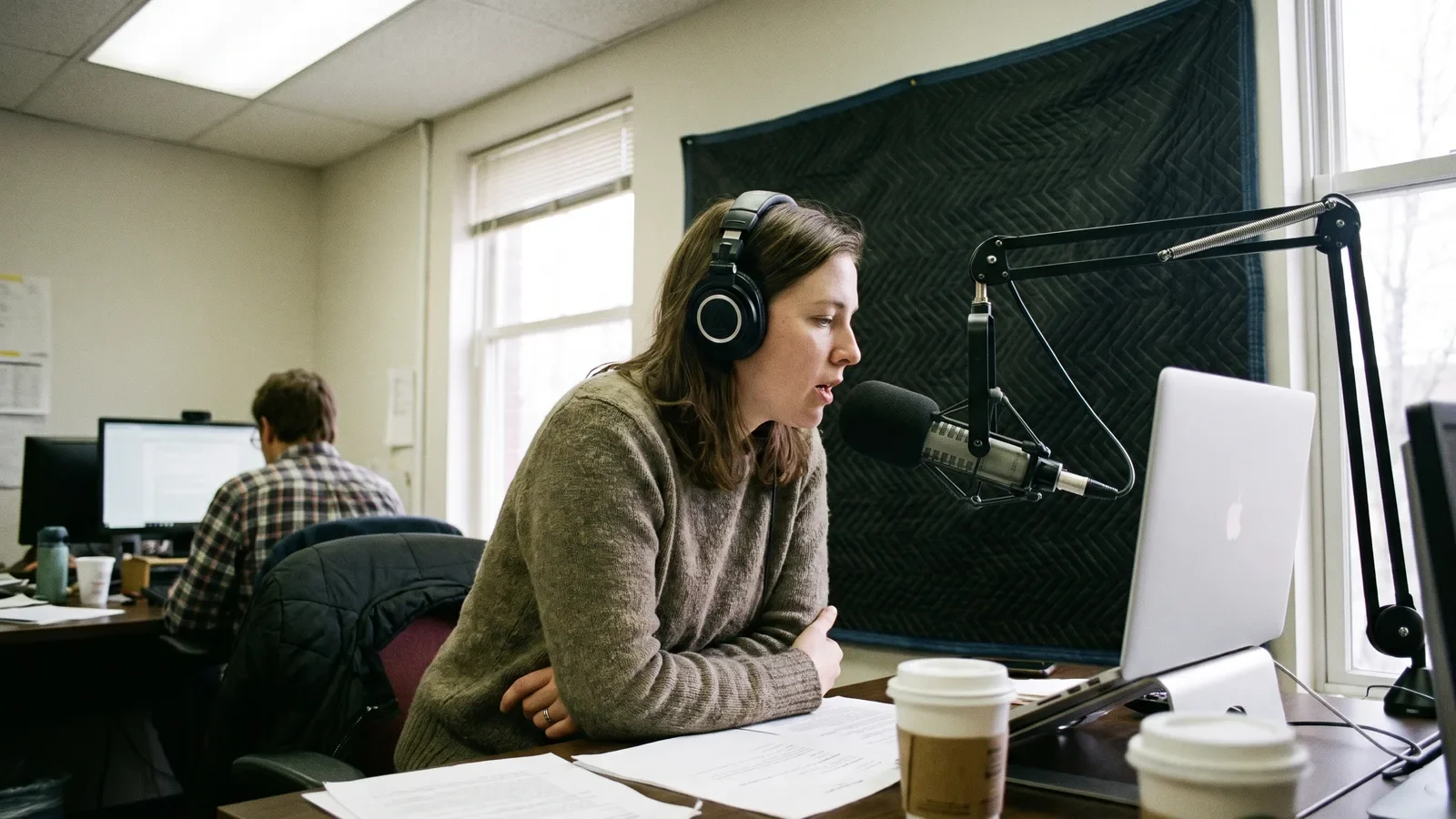 Reporter records voiceover into a desk boom mic with headphones, blanket pinned to wall behind her.